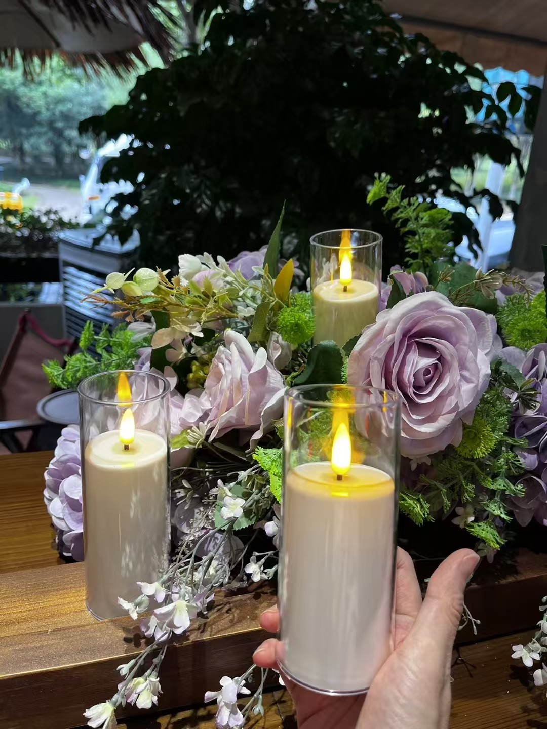 Glass-enclosed LED pillar candles displayed among pale purple flowers and greenery